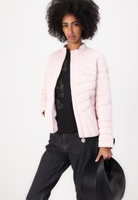 Light pink, quilted jacket with a high collar and two snap buttons. Worn over a black shirt and paired with dark jeans; holding a black handbag.