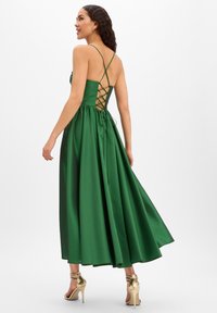 Grünes Satijn-Abendkleid mit ausgestelltem Rock und überkreuzter Schnürung am Rücken. Kombiniert mit goldenen, geschnürten High-Heel-Sandalen.