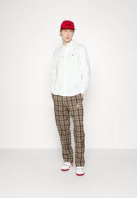 Chemise blanche à boutons, pantalon à carreaux brun avec broderies dorées, casquette rouge, chaussures blanches avec accents rouges, se tenant devant un fond uni.
