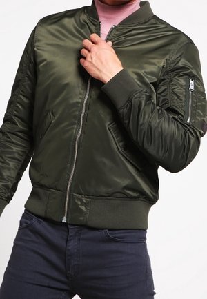 Personne portant une veste bomber vert foncé et un col roulé rose, ajustant la fermeture éclair de la veste avec la main droite.
