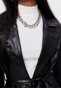 Czarna kurtka z imitacji skóry z wyraźnym kołnierzem, w połączeniu z białą bluzką o wysokim kołnierzu i masywnym srebrnym naszyjnikiem.