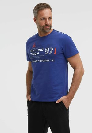 Man with short hair and beard wearing a blue "Sailing Tech 97" T-shirt with text and coordinates, hands in black pants pockets, looking right.