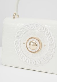 Sac à main en croûte de crocodile blanc embouti avec un fermoir circulaire en or présentant des accents tressés et des logos embossés, conçu avec une forme structurée et une poignée supérieure.