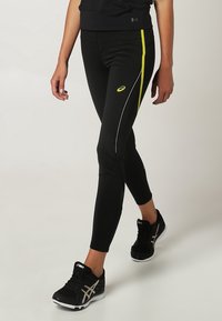 Černé sportovní legíny se žlutými a bílými bočními pruhy, s těsným střihem a materiálem odvádějícím vlhkost, doplněné černými teniskami.