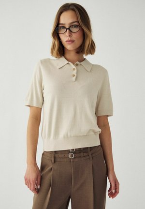 Femme aux cheveux mi-longs portant des lunettes noires, un polo beige à manches courtes boutonné et un pantalon marron taille haute avec double ceinture.