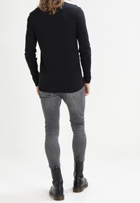 Osoba stojící zády, oblečená v černém dlouhém rukávu, šedých slim kalhotách a černých kotníkových botách, na bílém pozadí.