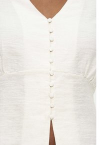 Κοντινό πλάνο λευκού υφασμάτινου φορέματος με υφή, με επτά κουμπιά καλυμμένα με ύφασμα που εκτείνονται κάθετα στον κεντρικό άξονα.