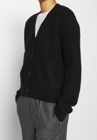 Černý cardigan z měkkého, jemného materi�álu. Má výstřih do V, tři knoflíky a přední kapsu. Volný střih s vroubkovaným lemem a manžetami.