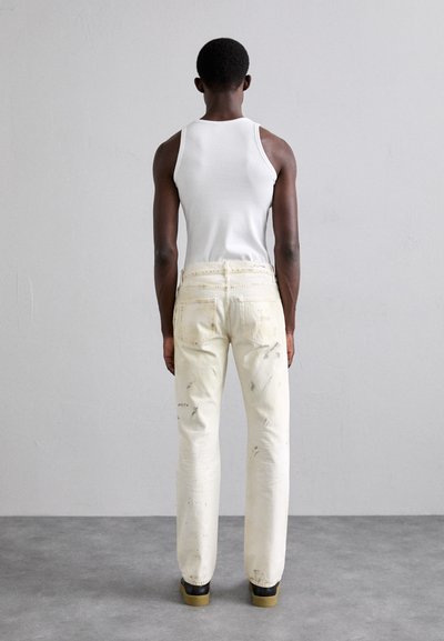 Débardeur blanc, jeans déchirés jaune clair avec des éclaboussures de peinture noire, coupe slim, associés à des chaussures noires à semelles en caoutchouc.