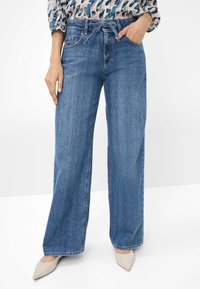 Wide-leg blue denim jeans with a high waist, front pockets, and a relaxed fit. Paired with beige pointed-toe heels.