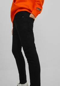 Černé džíny slim fit s předními kapsami a zapínáním na jeden knoflík, v kombinaci s jasně oranžovou mikinou.