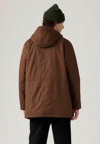 Brun isolerad jacka med huva, med en slät textur och en enda söm bak. Bärs över svarta byxor och kombineras med en grön stickad mössa.