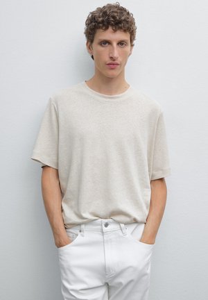 T-shirt di cotone beige con maniche corte, collo rotondo, vestibilità rilassata, indossata con pantaloni in denim bianchi, mani in tasca, su uno sfondo chiaro.