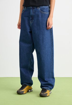 Osoba nosząca luźne granatowe dżinsy i żółto-czarne sneakersy stojąca na zielonej podłodze na tle białej ściany.