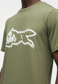 Λαδί βαμβακερό μπλουζάκι με λευκό γραφικό ψεκασμένο με σπρέι, που απεικονίζει ένα άλμα ζώο, με στρογγυλή λαιμόκοψη και κοντά μανίκια.