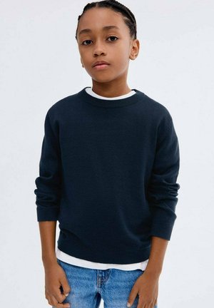 Marineblauer Strickpullover mit rundem Halsausschnitt, über einem weißen Hemd getragen, kombiniert mit blauen Jeans; hat lange Ärmel und eine weiche Textur.