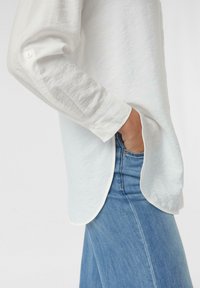 White textured long-sleeve shirt with buttoned cuffs, side slits, worn over blue denim, showing a relaxed fit and casual style.