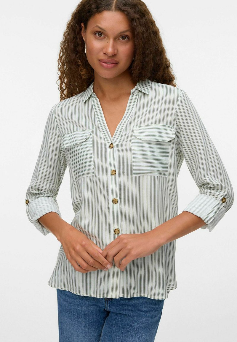Donna con capelli ricci che indossa una camicia bianca a bottoni con righe verticali verdi e jeans blu, mani delicatamente incrociate davanti.