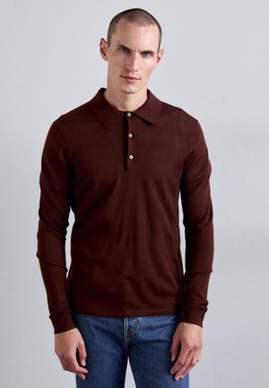Man die tegen een effen muur staat, gekleed in een donkerbruine polo met lange mouwen en een blauwe jeans, met een neutrale gezichtsuitdrukking.