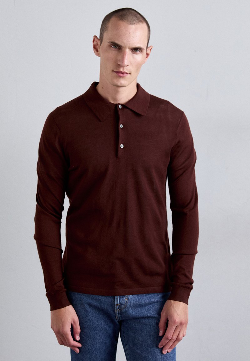 Mann steht an einer schlichten Wand, trägt ein dunkelbraunes langärmeliges Poloshirt und blaue Jeans, mit einem neutralen Gesichtsausdruck.