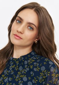 Navy blouse with a floral pattern featuring green leaves and white blossoms, textured fabric, pleated neckline, and gold earrings.