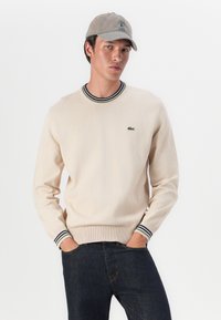 Bežový pletený sveter s rebrovanými manžetami, ktorý má námornícke a zelené pruhy a je ozdobený malým zeleným logom krokodíla na hrudi.