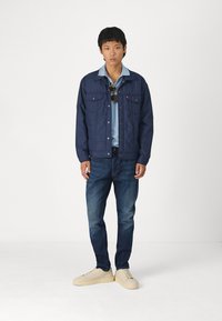 Marineblauwe gewatteerde jas met twee borstzakken, gedragen over een lichtblauwe overhemd met kraag, gecombineerd met donkere spijkerbroek en crèmekleurige sneakers.