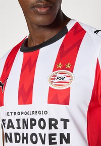 Raudonos ir baltos dryžuotos futbolo marškinėliai su juodu apykakliu, ant kurių yra PSV Eindhoven logotipas, dvi aukso žvaigždės ir juodas tekstas. Audinys su tekstūra.
