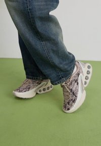 Snake-patterned sneakers in beige with a textured upper, white midsole, and visible air cushioning pods. Worn with blue denim jeans.
