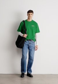 T-shirt de algodão verde sobre uma camisa de mangas longas branca, jeans azuis, sapatos de couro pretos e uma bolsa preta. Conjunto simples e casual com um corte estruturado.
