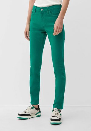 Personne portant un pantalon slim vert et des baskets blanches avec des accents noirs et verts, debout devant un fond clair uni.