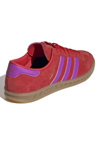 Czerwona zamszowa tenisówka z fioletowymi paskami, gumową podeszwą i sznurowanym designem, z złotym logo "Hamburg" i kontrastującymi przeszyciami.
