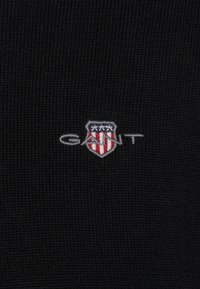 Tejido de punto negro con un pequeño logo bordado con un diseño de escudo, acentos en rojo y azul, y el texto "GANT" en gris.