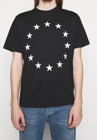 Černé bavlněné tričko s kulatým uspořádáním bílých hvězd potištěných na přední straně, s klasickým kulatým výstřihem a krátkými rukávy.