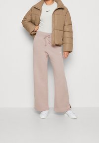 Brun pufferjacka över en ljusgrå t-shirt, kombinerad med rosa vida sweatpants med dragsko och vitt Nike-logga, avslutad med vita skor.