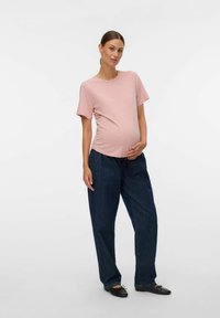 Gravid kvinna i en rosa kortärmad skjorta och lösa mörkblå byxor, stående och försiktigt hållande om sin mage, mot en enkel bakgrund.