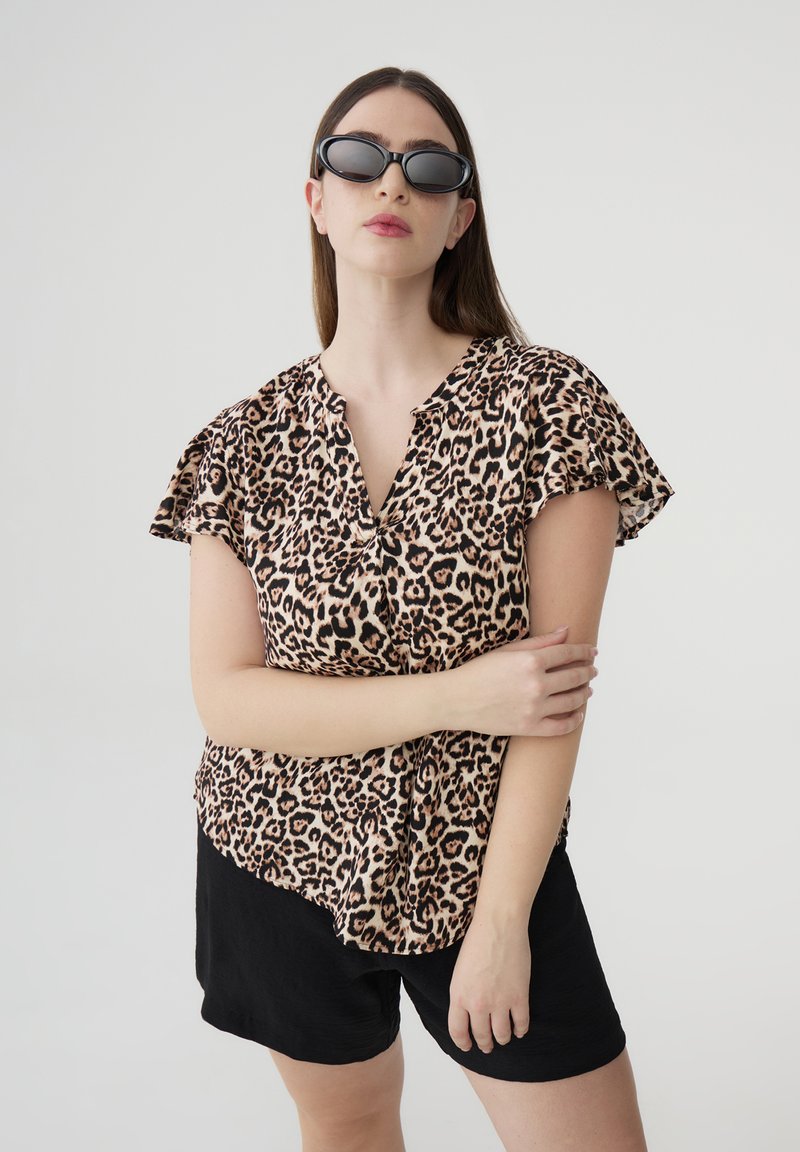 Blusa corta a maniche corte con stampa leopardata e scollo a V, realizzata in tessuto leggero. Il modello indossa pantaloni neri e occhiali da sole rettangolari.