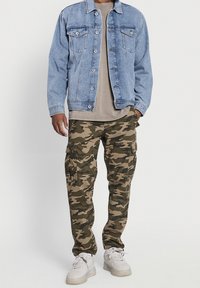 Lichtblauwe spijkerjack, beige t-shirt en camouflage cargobroek met zakken. Witte sneakers maken de outfit compleet.