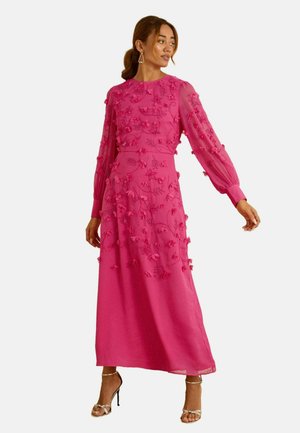 Žena v jasně růžových dlouhých maxi šatech s květinovými aplikacemi a stříbrnými vysokými podpatky pózuje s hlavou otočenou doprava.