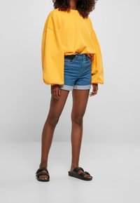 Sudadera amarilla de gran tamaño con hombros caídos, combinada con pantalones cortos vaqueros azules con dobladillo vuelto. Sandalias planas negras completan el conjunto.