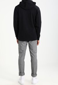 Persona che indossa un felpa nera con cappuccio, pantaloni grigi con risvolto, calzini bianchi e sneakers bianche, in piedi con la schiena rivolta su uno sfondo chiaro e semplice.