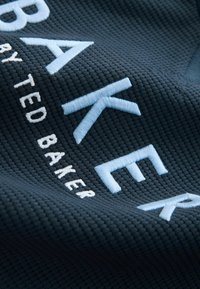Audums ar teksturētu tumši zilu audumu un paaugstinātu izšuvumu. Gaiši zilas burtus veido vārdi "BAKER" un "TED BAKER", kas kontrastē ar fonu.