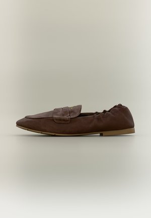 Brown flat loafer with pointed toe, elastic back, and decorative strap on a neutral background.