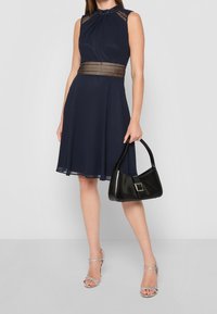 Námořnické modré bezrukávové šaty s přiléhavým živůtkem a rozšířenou sukní, zdobené dekorativním tkaným páskem; v kombinaci s černou kabelkou a stříbrnými podpatky.