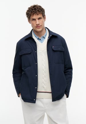 Ung mand med krøllet hår iført en navy jakke over en hvid kabelstrikket sweater og lyseblå skjorte med krave, stående med hænderne i lommerne.