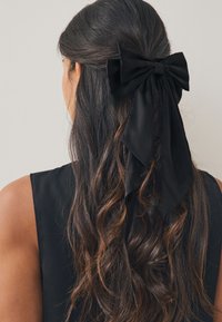 Zwarte satijnen haarlint gestyled in een grote strik, bevestigd aan golvend, donkerbruin haar. Het lint heeft lange, vloeiende uiteinden achter de strik.