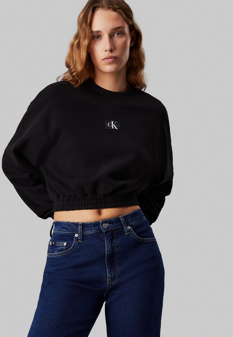 Felpa nera corta con cintura elastica, caratterizzata da un piccolo patch con logo CK. Abbinata a jeans blu scuro a vita alta.