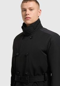 Čierny trench coat s dvojitou prednou časťou, veľkými gombíkmi, vysokým golierom a opaskom v páse; má elegantnú, hladkú tkaninu s matným povrchom.