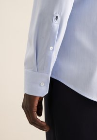 Camicia di un azzurro chiaro con texture, polsini esagonali, bottoni bianchi e un dettaglio floreale ricamato di colore nero sulla manica. Tessuto liscio.