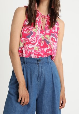 Frau trägt eine rosa geblümte ärmellose Bluse mit Schleifenkragen und hoch taillierte blaue Hosen mit weitem Bein vor einfarbigem Hintergrund.